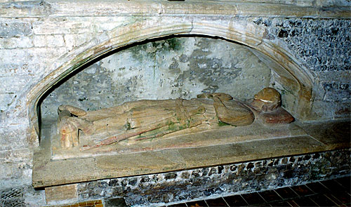 In a tomb recess in the chapel is this effigy of a man clad in a long military style surcoat, tippet and hood. Feet spurred and resting on a dog or a lion. At left side a dagger and a sword slung from belt buckled over hips. Late 13th century but the face has been restored at some later date.